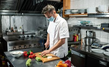 Professional young chef in uniform and protective mask bending over table in a ghost kitchen while chopping fresh zucchini and other vegetables